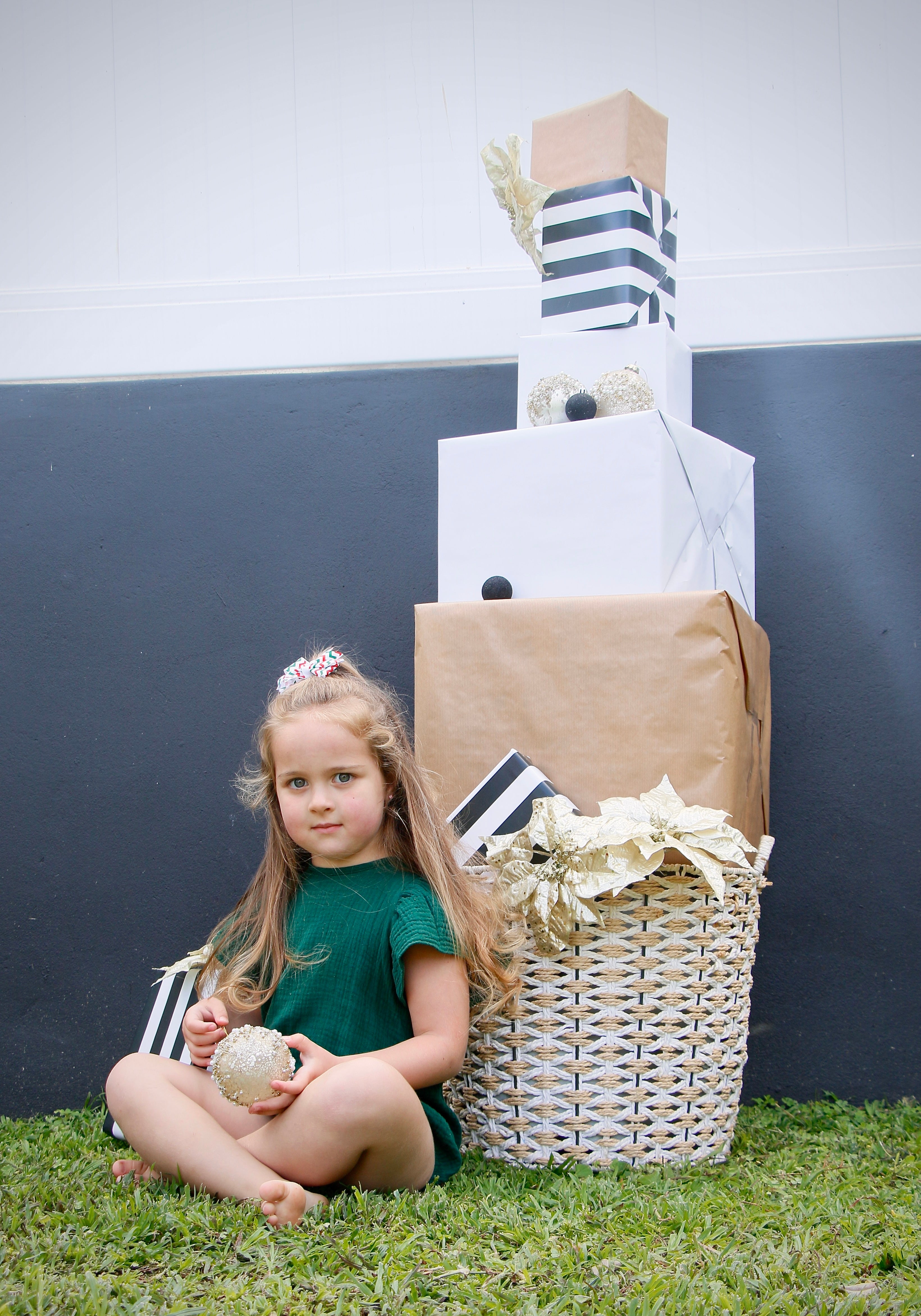 Young girl sitting on grass next to a large gift basket with wrapped presents against a plain background